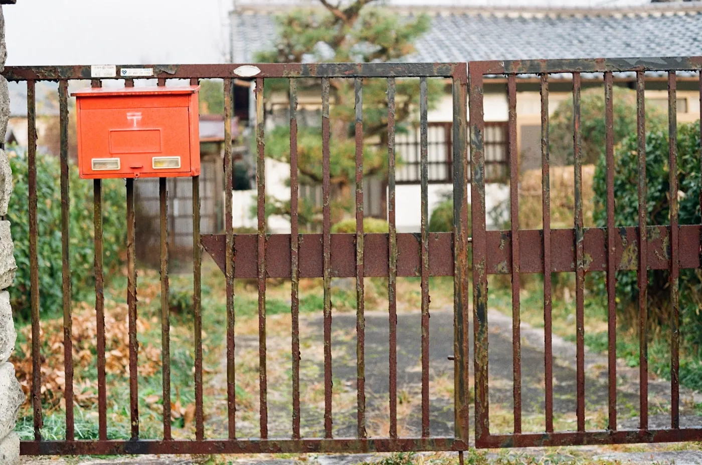さびた門扉に取り付けられた赤い郵便受けと、奥に見える住宅の庭