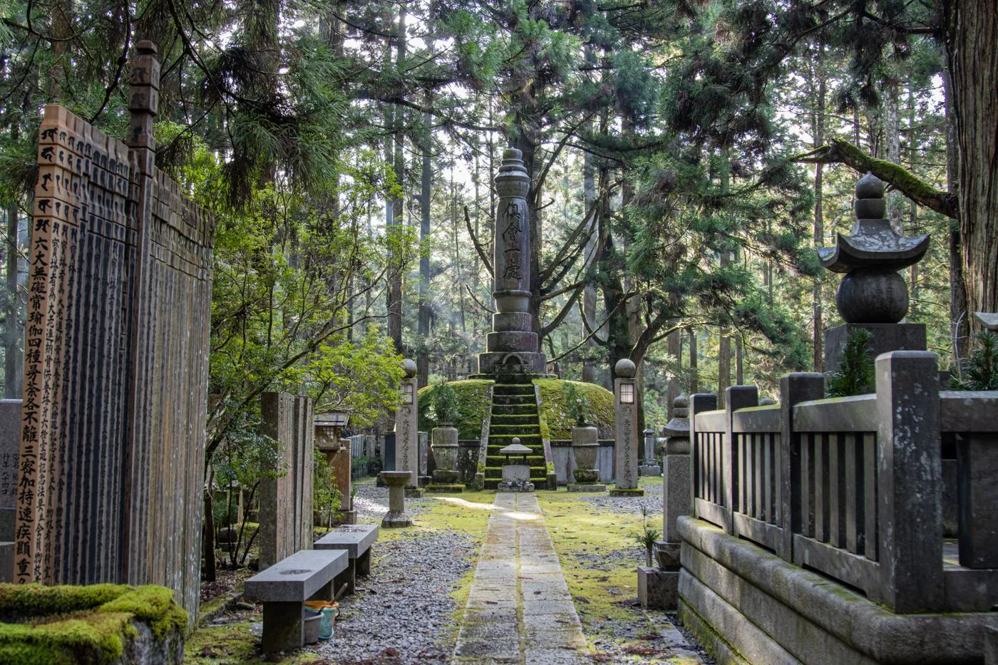 杉木立に囲まれた墓地の石畳の参道。奥に文字の刻まれた石塔が立ち、両側に石灯籠と玉垣が並ぶ