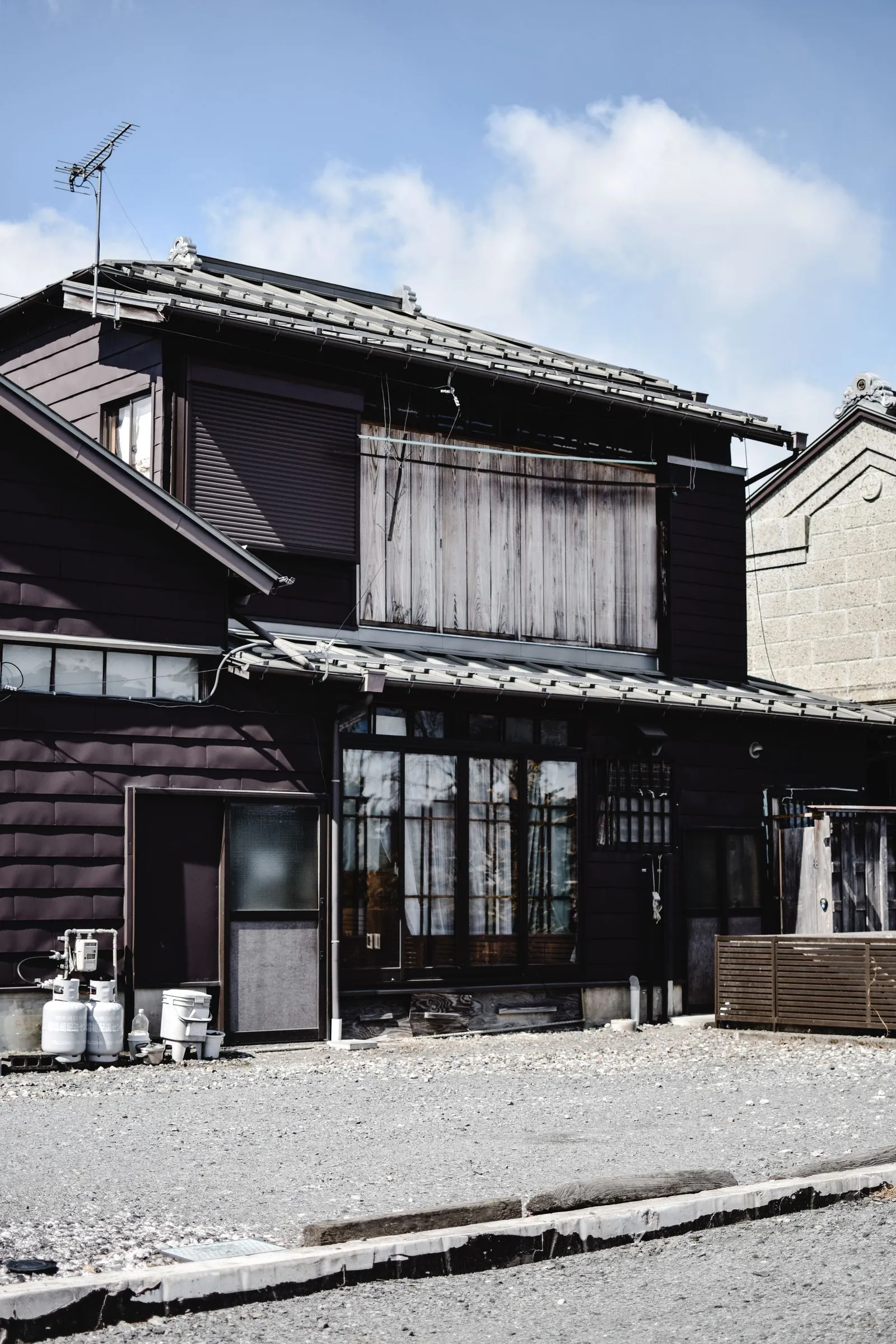 雨戸を閉めた黒い板張りの二階建て日本家屋と、外壁沿いに置かれたガスボンベ