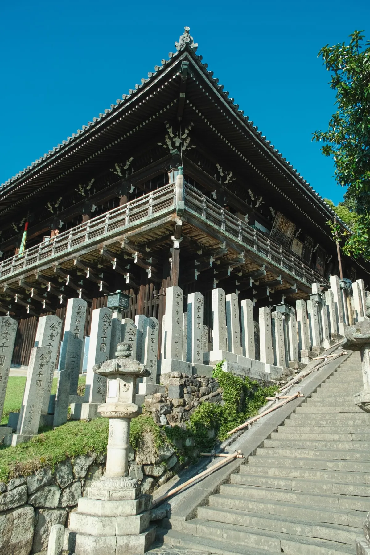 寺院の木造建築の軒と、両側に石柱と石灯籠が並ぶ石段