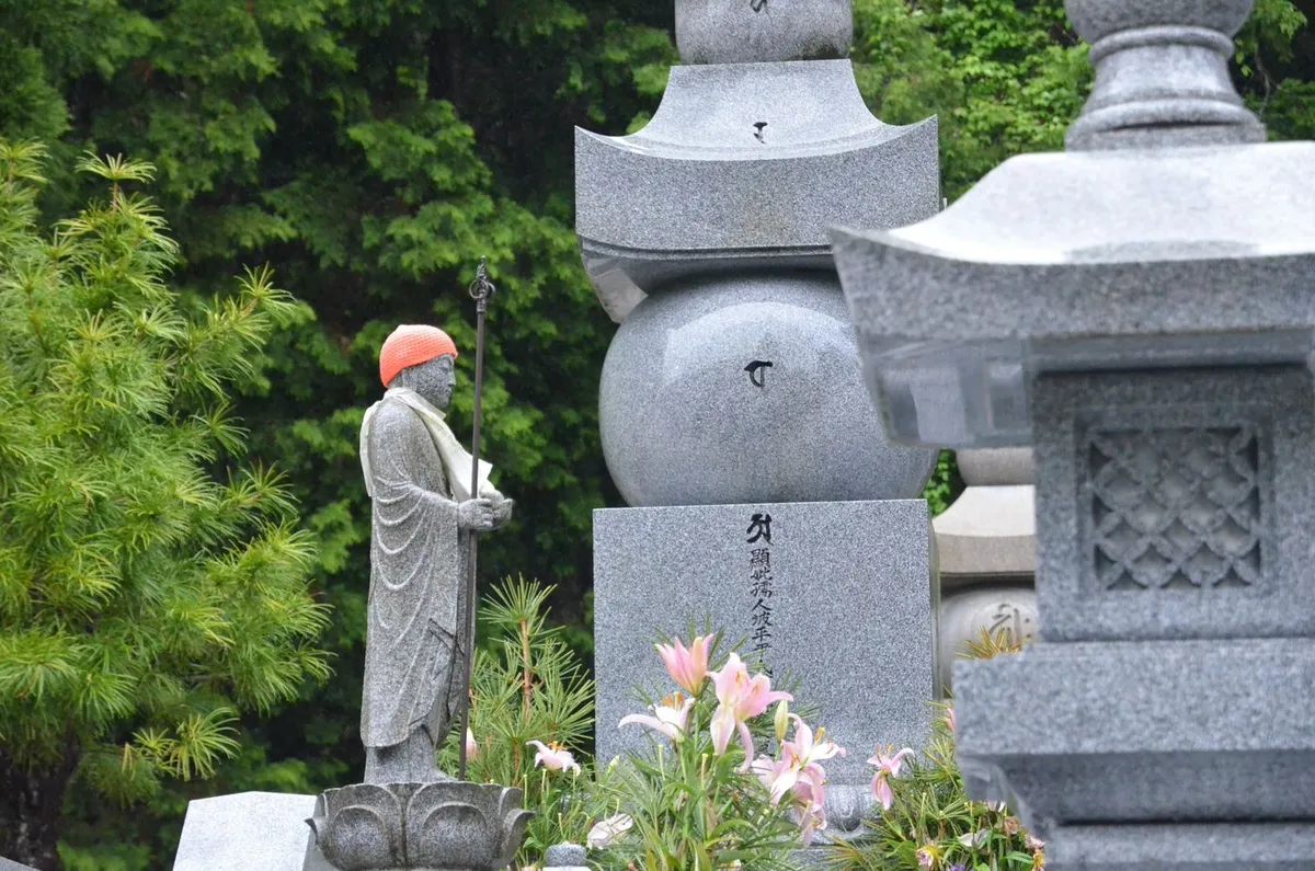 赤い帽子と衣をまとった石像のそばに、石塔と石灯籠、ユリの花が並ぶ