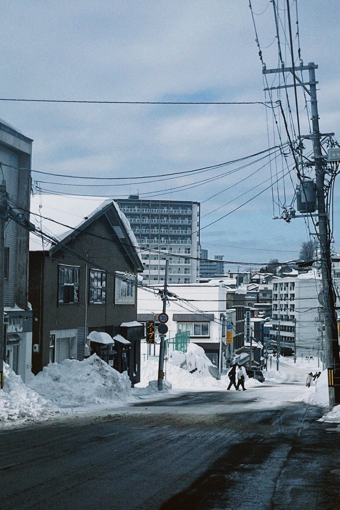 雪に覆われた日本の街並み。曇り空の下に建物と電線が見える