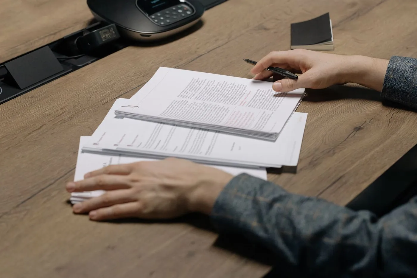 木製の机に置かれた書類の束にペンを持った手を添えている様子